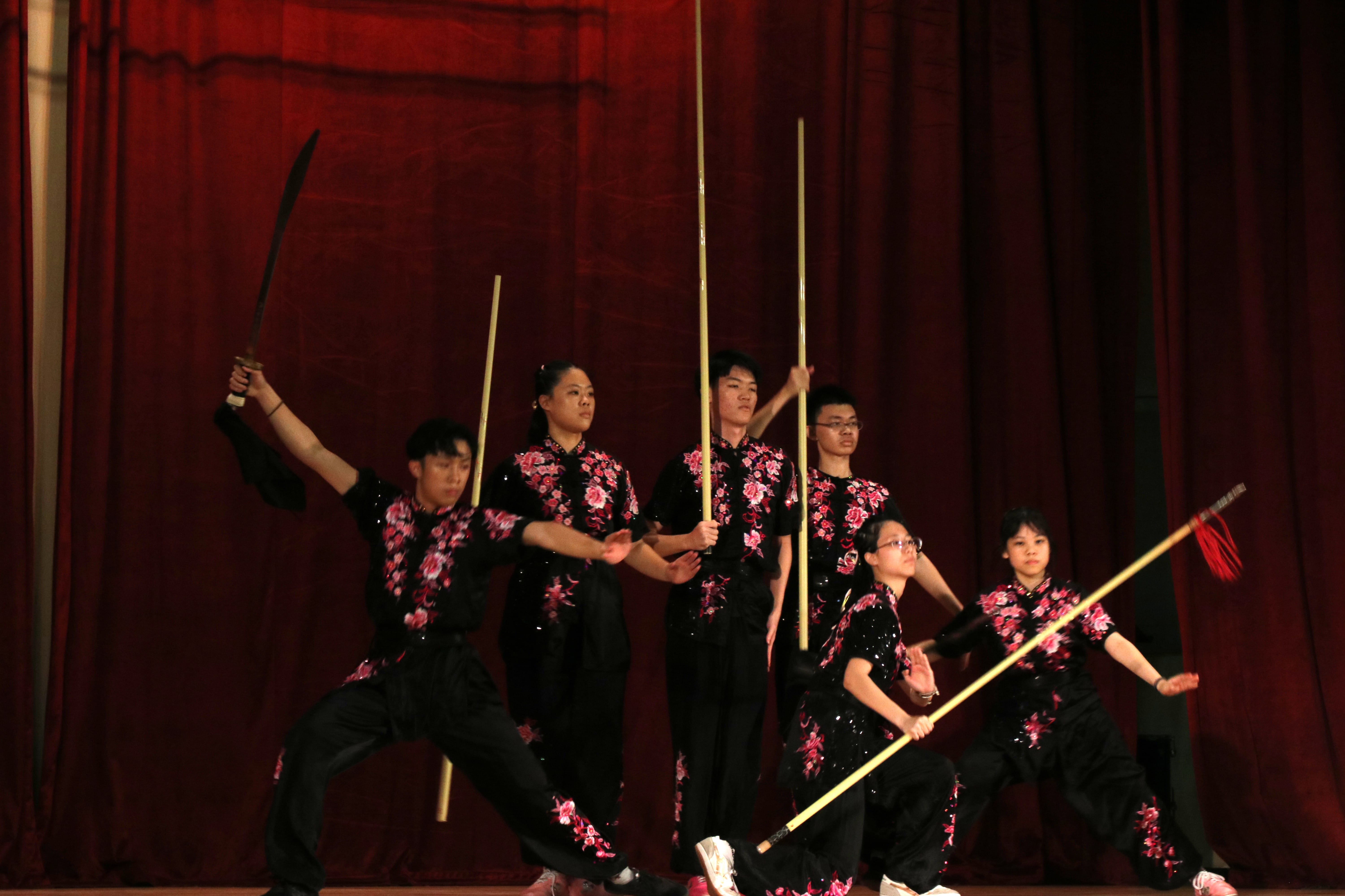 Six TMJC wushu students in black and pink floral uniforms perform a weapons routine on stage during an Open House showcase, wielding staffs and a sword against a red curtain backdrop